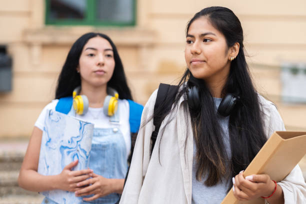 istock Two hispanic students walking outside a school building with headphones and backpacks, carrying folders 2169219416