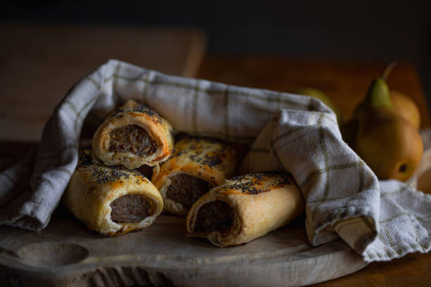 A close-up shot of four sausage rolls stacked on top of one another on a plate. They are covered with a serving towel and a teapot can be seen in the background.\n\nVideos are available similar to this scenario.