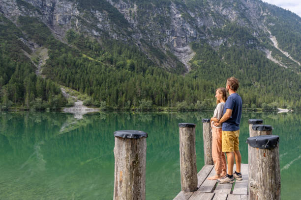A couple stands close, embracing on a wooden dock by a serene, reflective lake, surrounded by lush mountains. The tranquil setting emphasizes connection with nature and each other.