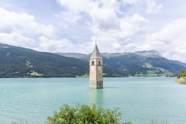 The iconic bell tower of a submerged church rises from the turquoise waters of Lake Resia, set against a backdrop of lush mountains under a partly cloudy sky.