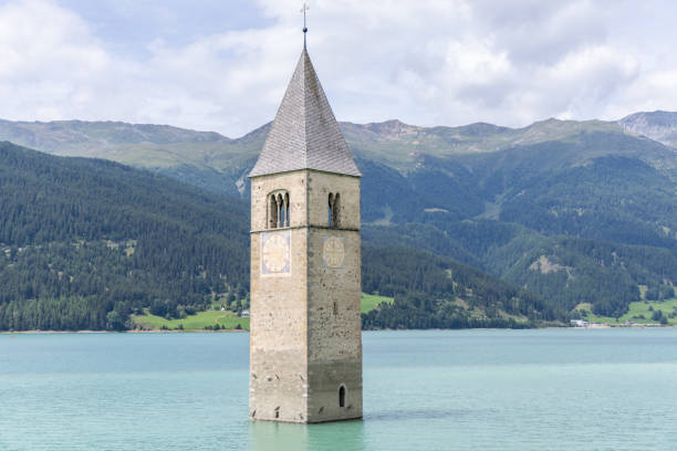 The iconic bell tower of a submerged church rises from the turquoise waters of Lake Resia, set against a backdrop of lush mountains under a partly cloudy sky.