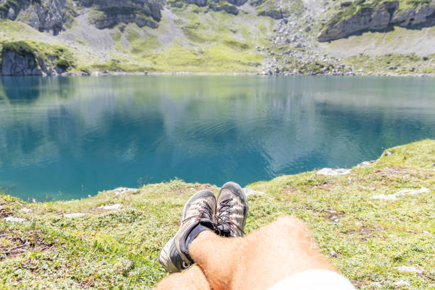 A person relaxes by a calm mountain lake, with their hiking shoes in the foreground, enjoying the peaceful surroundings and clear, blue water on a sunny day.