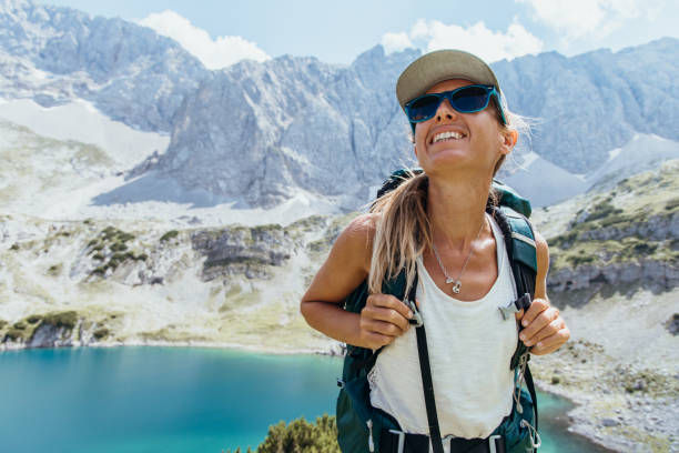 A happy female hiker with a backpack smiles as she takes in the stunning view of a turquoise mountain lake and towering rocky peaks on a bright, sunny day.