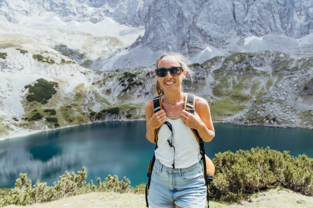 A cheerful female hiker with a backpack smiles as she enjoys the stunning view of a tranquil mountain lake and rugged peaks in the background, capturing the joy of outdoor adventure.