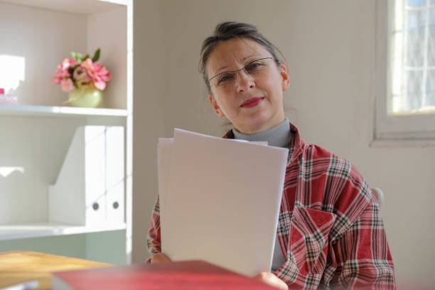 An elderly woman, wearing a plaid shirt and glasses, is captured smiling while reading through documents at home. The bright, cozy environment suggests themes of financial planning, retirement, or personal growth, making this image versatile for various senior-related content.