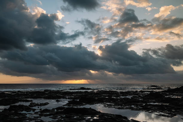 The sky is cloudy and the sun is setting. The ocean is calm and the rocks are visible