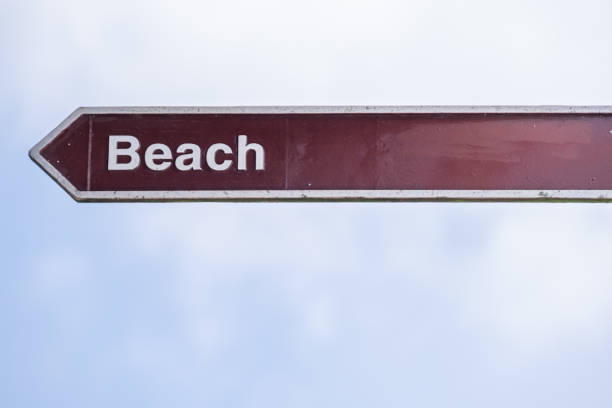 Beach sign, thin, isolated, brown maroon colour, white text lettering, pointing left - isolated against light cloudy sky.