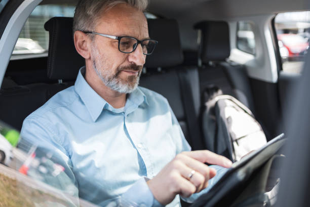 istock Business commuter using a tablet to work remotely while riding in a taxi. Mature man with glasses and a serious expression, focused on his work. Concept of remote work and commuting. 2169359563