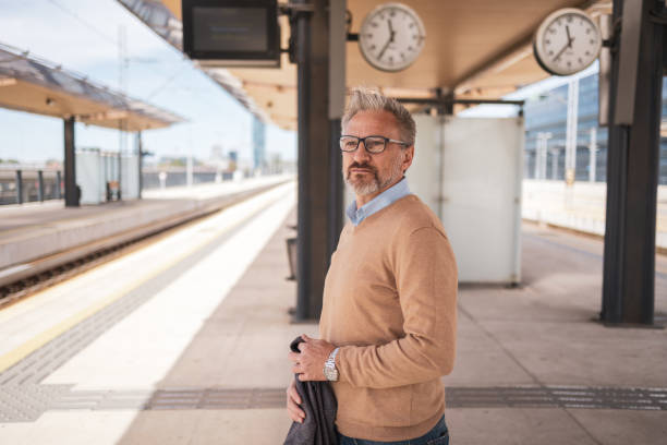 istock Mature man standing at a modern train station platform, looking confident and thoughtful. Wearing glasses and casual attire, he appears ready for a journey or daily commute, embodying a professional yet relaxed demeanor. 2169359569