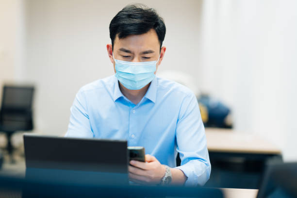 istock Asian business man in protective face mask using smart phone and computer in office. 2169367067