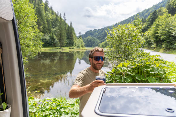 A man places a cup of coffee on the counter of his camper van, with a beautiful lake and lush forest in the background. Embracing the serenity of nature.