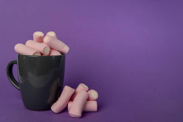 Detailed horizontal photo of a gray cup filled with marshmallows, with a small mound of marshmallows positioned to the left. The scene is set against a striking purple background