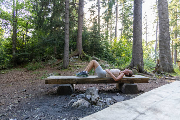 A woman lies on her back on a wooden bench in a peaceful forest, resting after a workout. The serene natural environment contributes to her relaxation and recovery.