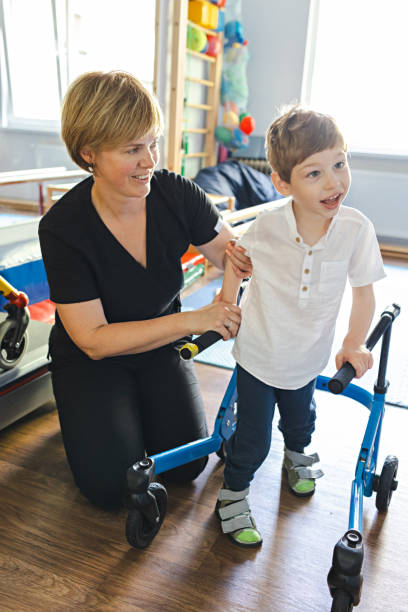 Handsome young boy with movement disorders practices walking with support from a therapist in rehabilitation clinic