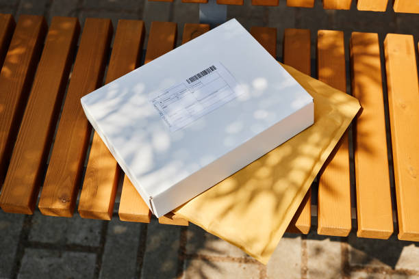 Mail package and envelope placed on wooden bench in outdoor setting being partially shaded by sunlight filtering through leaves