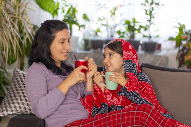 A cozy moment as mother and daughter snuggle under a red blanket, sipping tea and chatting on the sofa.