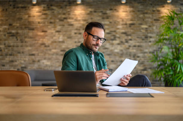 istock Focused male accountant in eyeglasses reading documents while working over laptop at desk in office 2169448148