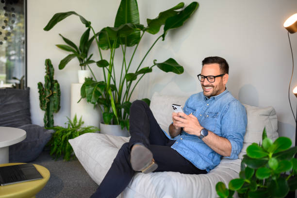 istock Handsome manager in glasses texting over mobile phone while relaxing on sofa at his modern workplace 2169448328