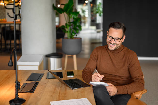 istock High angle view of smiling businessman writing notes while working over laptop at desk in office 2169448359