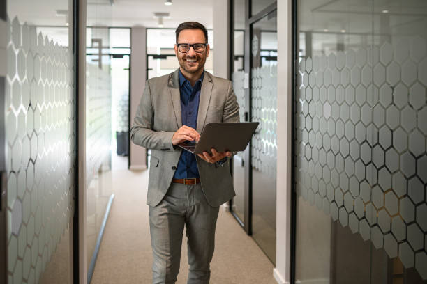 istock Confident male professional working over laptop and smiling at camera while standing in office corridor 2169448384