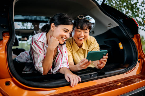 istock Two smiling female friends using smart phone while laying in car trunk together 2169451030