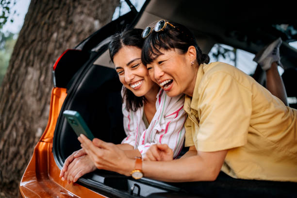 istock Two smiling female friends using smart phone while laying in car trunk together 2169451072