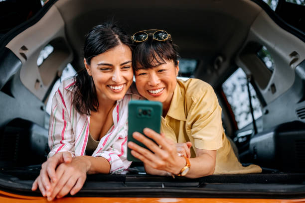 istock Two smiling female friends using smart phone while laying in car trunk together 2169451083
