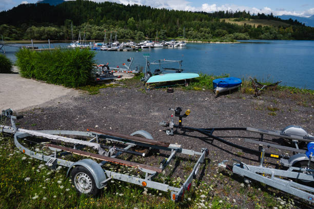 The image features a collection of boat trailers and small boats on a gravel shore near a lakeside marina. In the background, yachts are docked on the calm water, surrounded by lush green hills and a forested area under a partly cloudy sky.