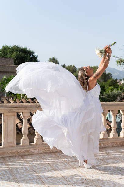 Bride is standing on a terrace, raising her bouquet while her long dress flows in the wind