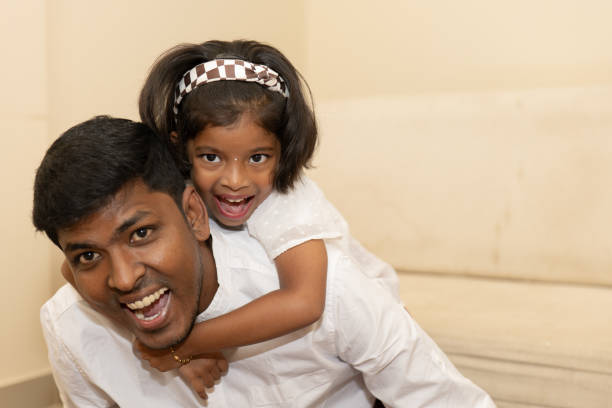 A father is playfully engaging with his daughter, who is sitting on his back and smiling brightly, showcasing their loving bond in a warm home environment.