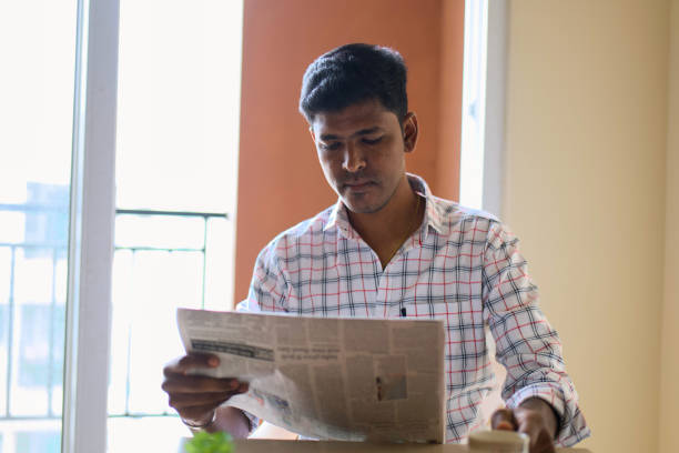 This image is about Man reading a newspaper while sitting at a table near a window in a cozy indoor space during daytime