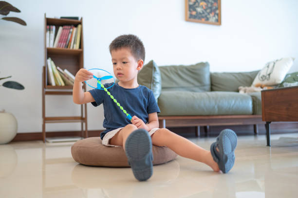 A young boy in a blue T-shirt and beige shorts is seated on a round brown cushion on a smooth, shiny floor in a living room, focused on putting together a blue flying propeller toy