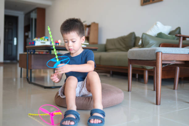 A little boy in a blue shirt and shorts is sitting on a round brown cushion on a shiny floor in a well-furnished living room, focused on assembling a colorful flying propeller toy