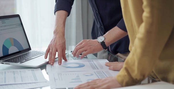 istock Business people, professionals analyzing financial charts and data on a desk while standing and using laptop computer in office. Dark blue shirt and yellow blouse 2169490393