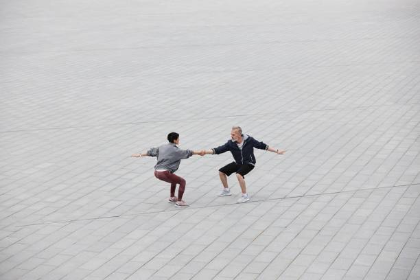 Wide angle shot of fit senior man and woman doing partner exercise balancing in seated position holding hands in middle of large tiled gray square at city park, copy space