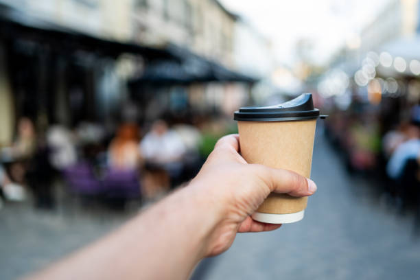 istock hand holding a disposable coffee cup with a black lid, set against a blurred urban background with people walking. focus is on the cup, highlighting concept of takeaway coffee in a city environment. 2169497063