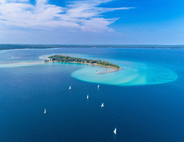 A view from above of the Higgins Lake Lightning Regatta Race. A small island with lush greenery is surrounded by vibrant blue and turquoise waters. Several sailboats are scattered across the water, with a partly cloudy sky overhead. Higgins Lake, Michigan. 7/28/24