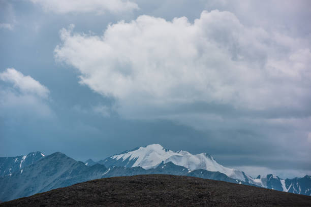 Dramatic layer view to large wide snow-capped mountain range far away, big rocky ridge silhouette and stony rounded hill in center close up under rainy gray sky. Overcast scenery in high mountains.