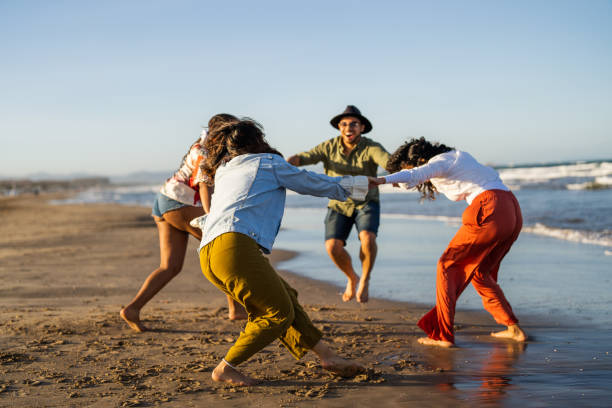 A group of diverse friends in casual beachwear joyfully dance on the shore, their bare feet sinking into the wet sand as waves gently break in the background.