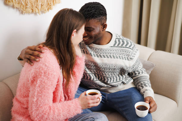 istock Smiling multiethnic couple hugging while drinking coffee on couch at home 2169559104