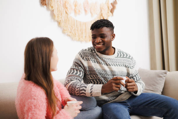 istock Smiling african american man holding cup while talking to girlfriend on couch at home 2169559105