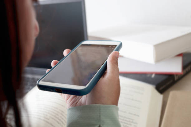 istock Hand holding smartphone and using AI ​​translation application while reading textbook  in the library. Woman student learning foreign language online on mobile phone. 2169567303