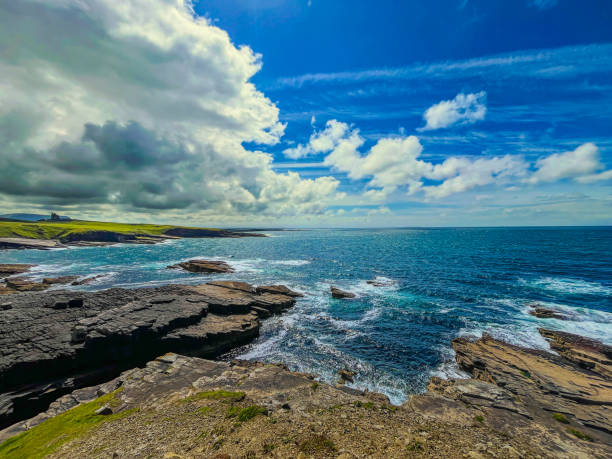 A captivating view of the rugged coastal cliffs at Mullaghmore in County Sligo, Ireland. The scene captures the vibrant blue Atlantic Ocean meeting dramatic rocky formations under a sky filled with dynamic cloud patterns. A distant castle on the coastline adds to the landscape’s mystique, illustrating the natural beauty and historical depth of this iconic Irish location.