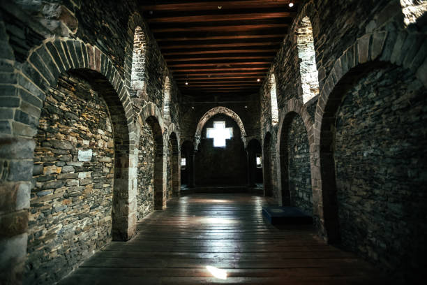 This photograph captures the interior of a historic stone building with a series of arched doorways and a central cross-shaped window at the far end. The stone walls, wooden beams on the ceiling, and wooden floorboards create a rustic, medieval atmosphere. The soft natural light entering through the small windows and the cross-shaped window at the back adds a contemplative mood to the space.
