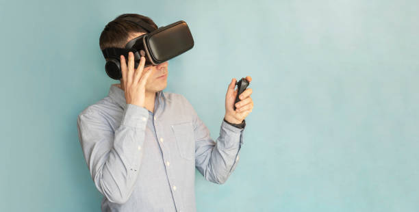 istock Young smiling man in VR glasses playing video game with joystick on a blue background, siting on the couch while having virtual reality experience, copy space 2169658279