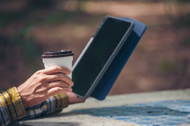 Person holding tablet and disposable coffee cup outdoors. Man hand using phone to learn online at home office. Casual outdoor work or study concept.