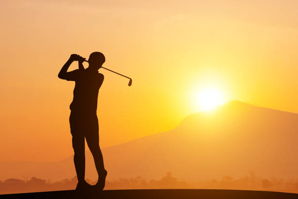 Golfers' hit golf ball toward the hole at sunset silhouetted. Golden morning sky in winter, misty high mountain background.