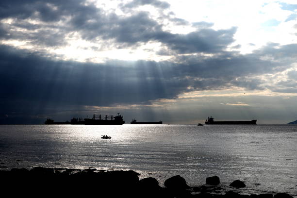This dramatic photograph captures the interplay of light and shadow over the waters near Stanley Park, Vancouver. Sun rays pierce through thick clouds, casting a luminous glow across the sea, while large ships are silhouetted against the horizon. In the foreground, the calm water reflects the light, creating a tranquil yet powerful scene. The solitary kayak further emphasizes the vastness of the ocean and the grandeur of nature, contrasting with the industrial presence of the ships. This image beautifully conveys the serene yet dynamic atmosphere of Vancouver's coastal landscape.