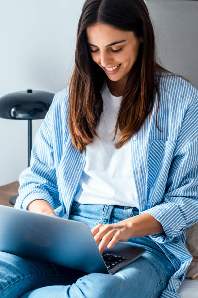 istock Young woman sitting on her bed, smiling while chatting online with friends using a laptop and wireless internet. A passionate technology enthusiast, she balances remote work, socializing, and modern digital communication 2169685976