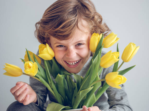 Excited boy in casual wear looking at camera with smile near bouquet of colorful tulips on white background in studio